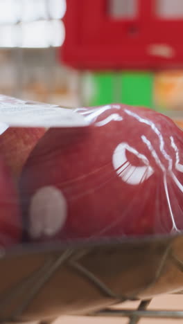 una mano colocando un paquete de manzanas en un carrito de compras, capturado en un entorno dinámico de un centro comercial. esta vista inmersiva destaca el acto de comprar comestibles con una cámara de mano