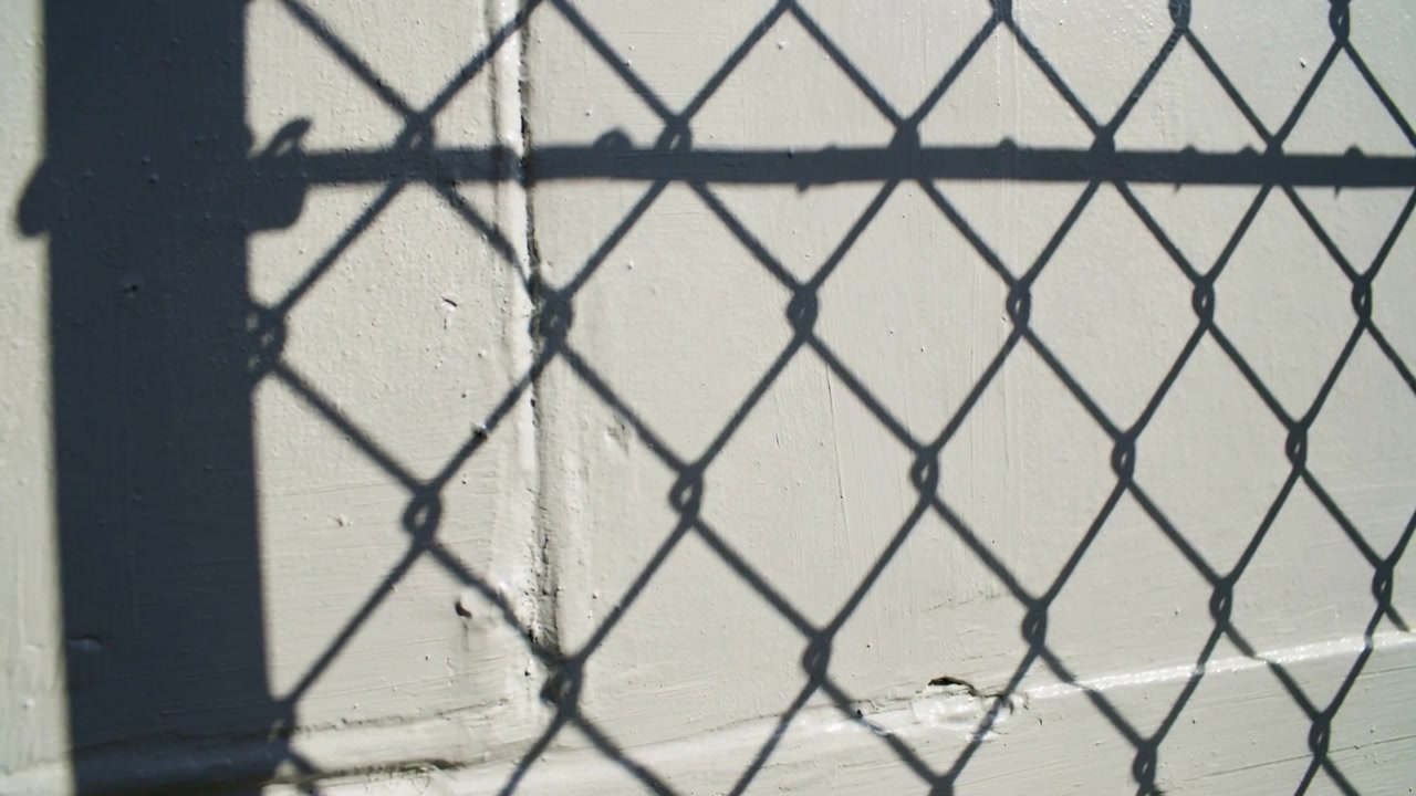 Sol brillante golpeando una valla de tela metálica proyectando una sombra de malla en una pared de hormigón pintada, sombras de postes de barandilla