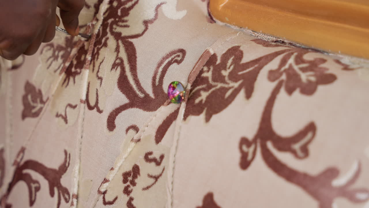 Upholstery repair closeup showing hands securing decorative tack into patterned fabric, upholsterer attaching trim to wooden frame, bead and stud embellishment, careful handwork with needle