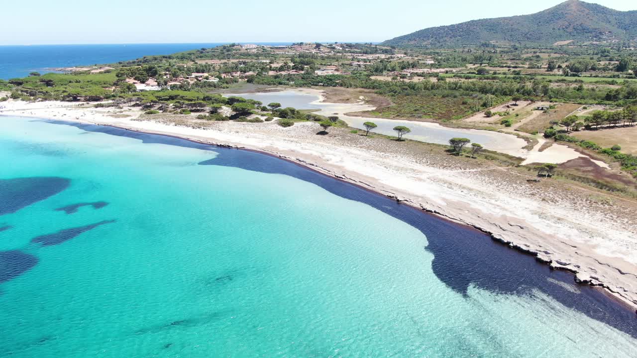 This video follows a forward motion  giving view of a crystal clear sea line with white sandy shores near a tree line forest. Parallel to the sea coastline is a small water pond