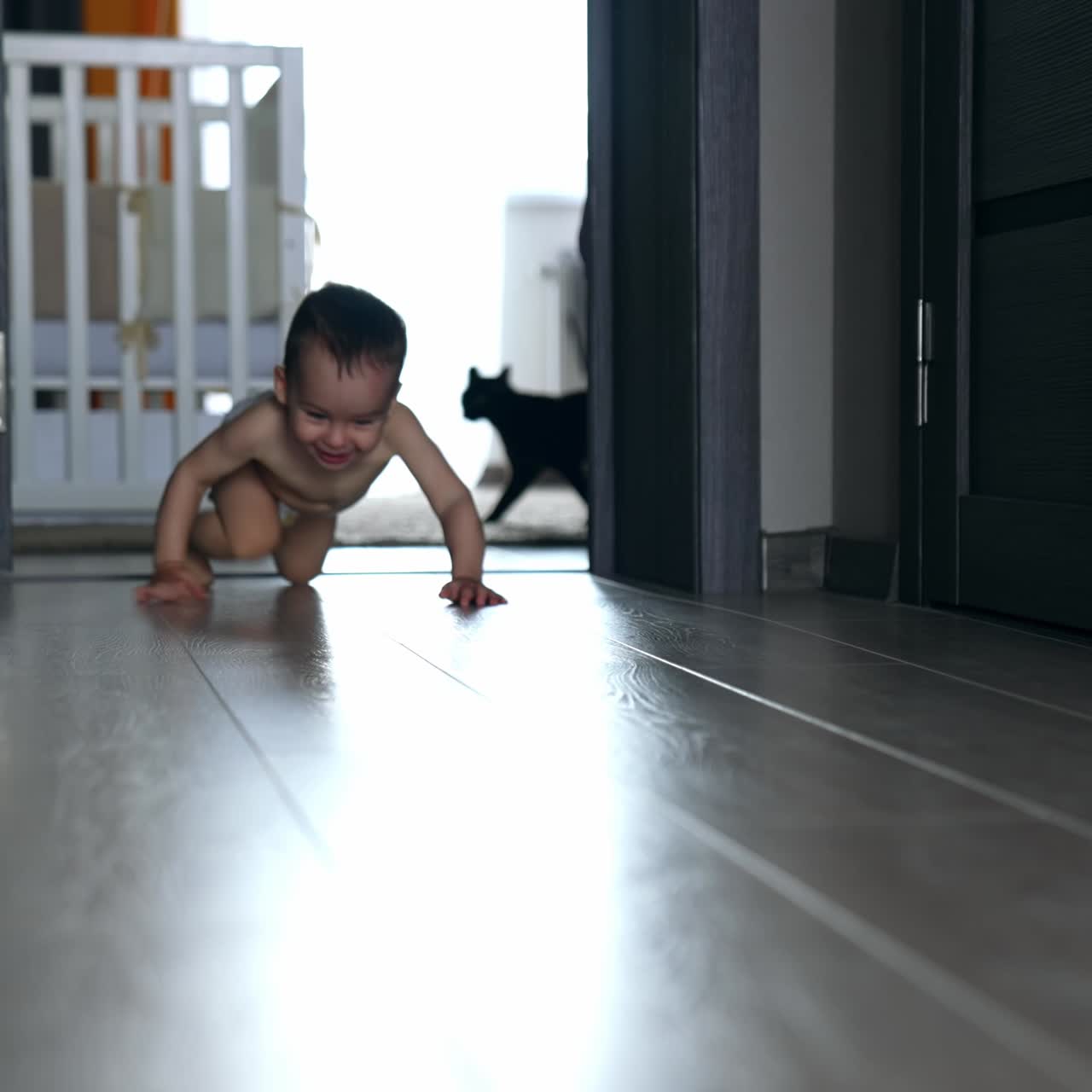 Happy smiling adorable baby in diaper crawls by the room. Black cat follows the child