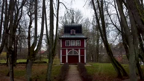 Aerial-drone-backing-up-from-a-small-red-chapel-between-barren-forest-trees-in-autumn