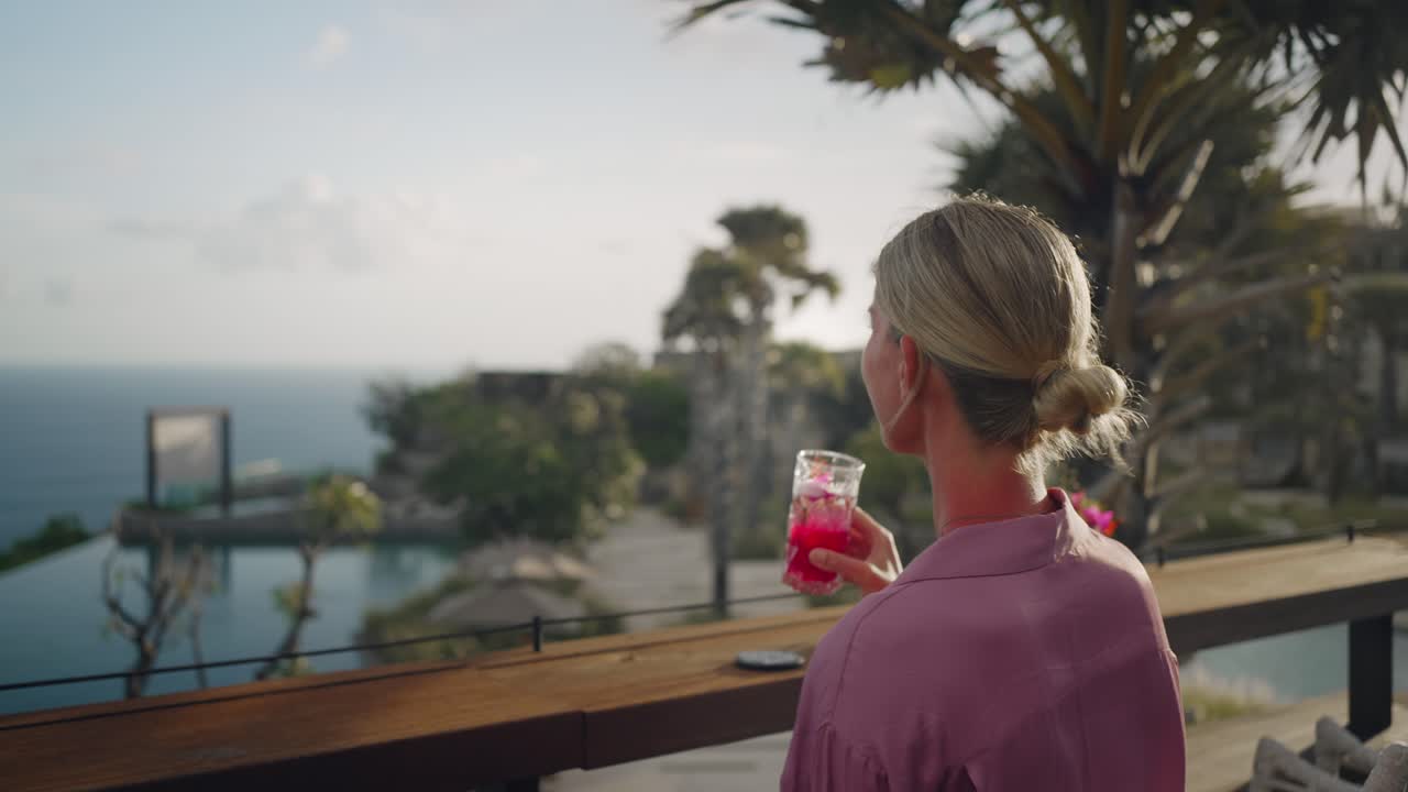 Woman grabs tropical drink from table, sipping cocktail and enjoys ocean view
