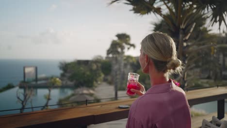 woman grabs tropical drink from table, sipping cocktail and enjoys ocean view