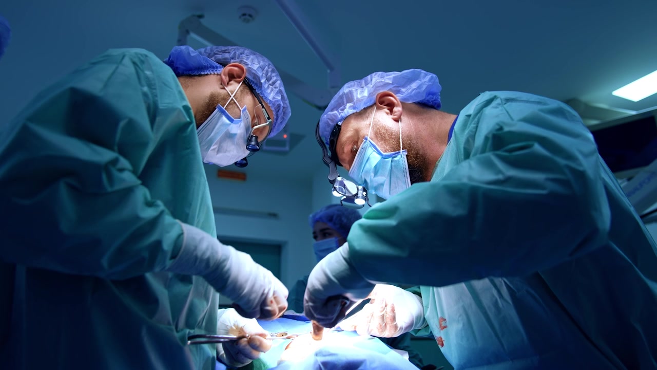 Chirurghi caucasici barbuti eseguono un intervento maxillo-facciale. Un'infermiera strumentista si trova sullo sfondo. Vista dal basso