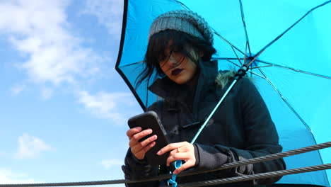 Atractiva-Mujer-Joven-Enviando-Mensajes-De-Texto-En-Un-Teléfono-Inteligente-Sosteniendo-Un-Paraguas-Bajo-Un-Cielo-Azul-Mientras-Pasan-Las-Nubes-De-Tormenta