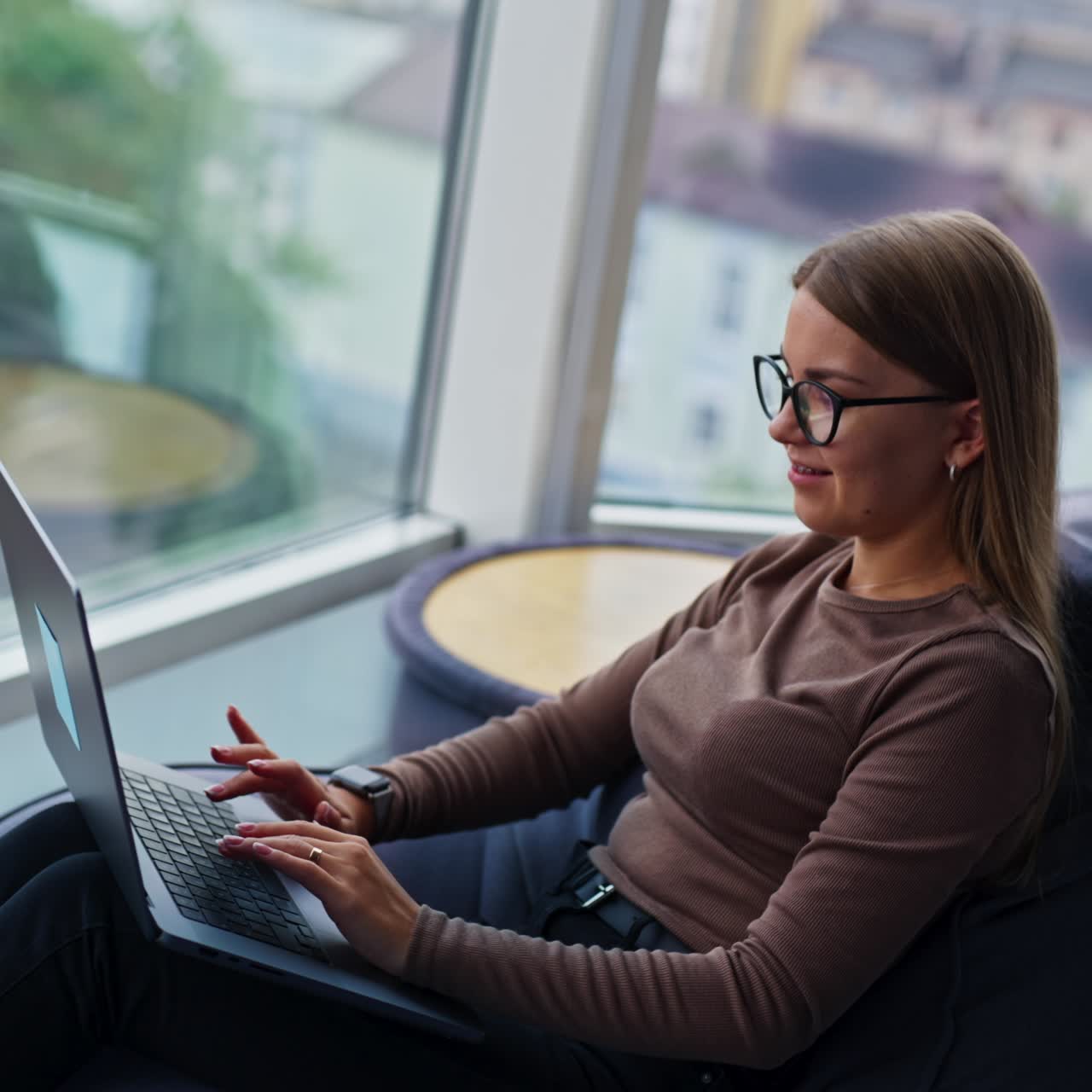 Woman in glasses opens laptop. Female starts typing on her computer sitting in comfortable bean bag chair. Side view. Blurred windows at backdrop