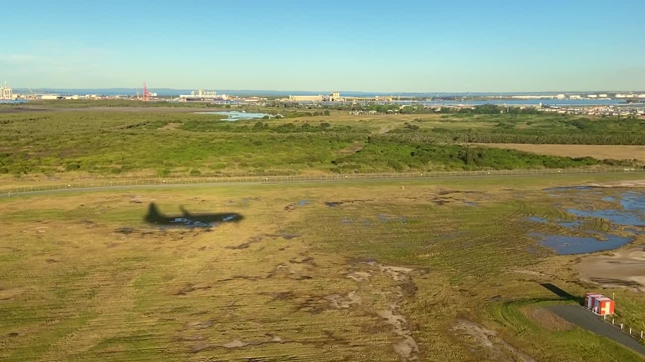 Low aerial shot with shadow of landing airplane, over mangroves and marshlands with port infrastructure, waterways and coastal pools, as plane lands smoothly on the runway