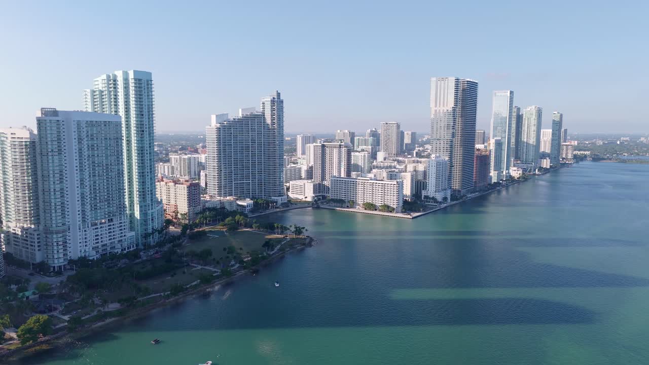 Aerial drone view of Miami’s downtown waterfront with modern high-rise buildings, a city park, and the calm turquoise waters of Biscayne Bay under a clear sky.