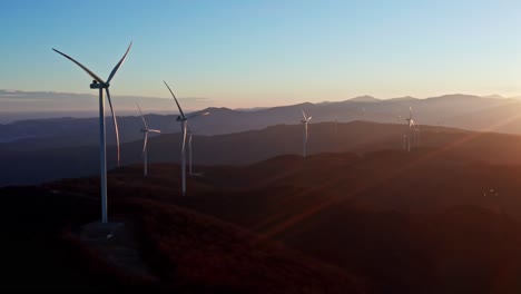 Toma-Aérea-Temprano-En-La-Mañana-De-Turbinas-Eólicas-En-Un-Terreno-Montañoso-Con-El-Amanecer-Proyectando-Una-Luz-Dorada