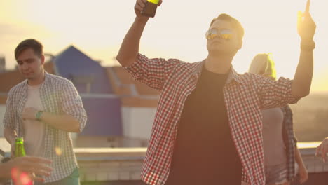 a young male in trendy glasses with beer moves in a dance on the party with his friends on the roof. he moves his hands in dance