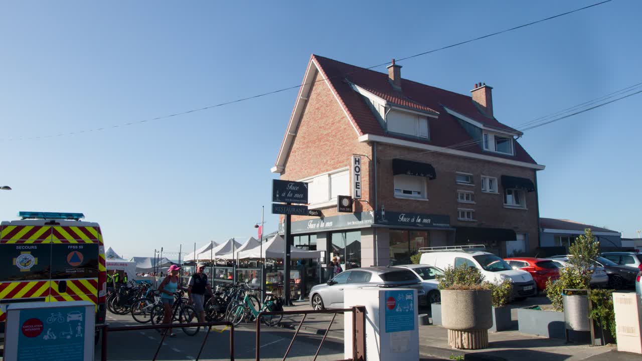 Cyclist rides past parked cars, bicycles, and market tents on sunny day in coastal town