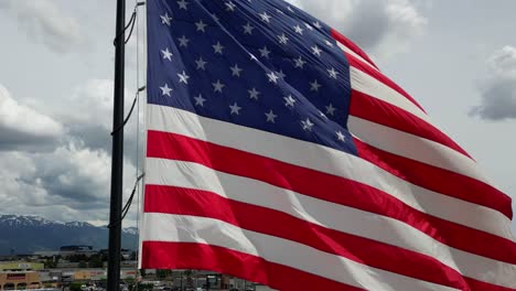 Die-Amerikanische-Flagge-Weht-An-Einem-Schönen-Sonnigen-Sommertag-Im-Wind-Mit-Vielen-Weißen-Wolken-Und-Blauem-Himmel-Mit-Blick-Auf-Die-Wasatch-Berge,-Während-Die-Drohne-Um-Den-Fahnenmast-Fliegt-–-In-4K-60fps