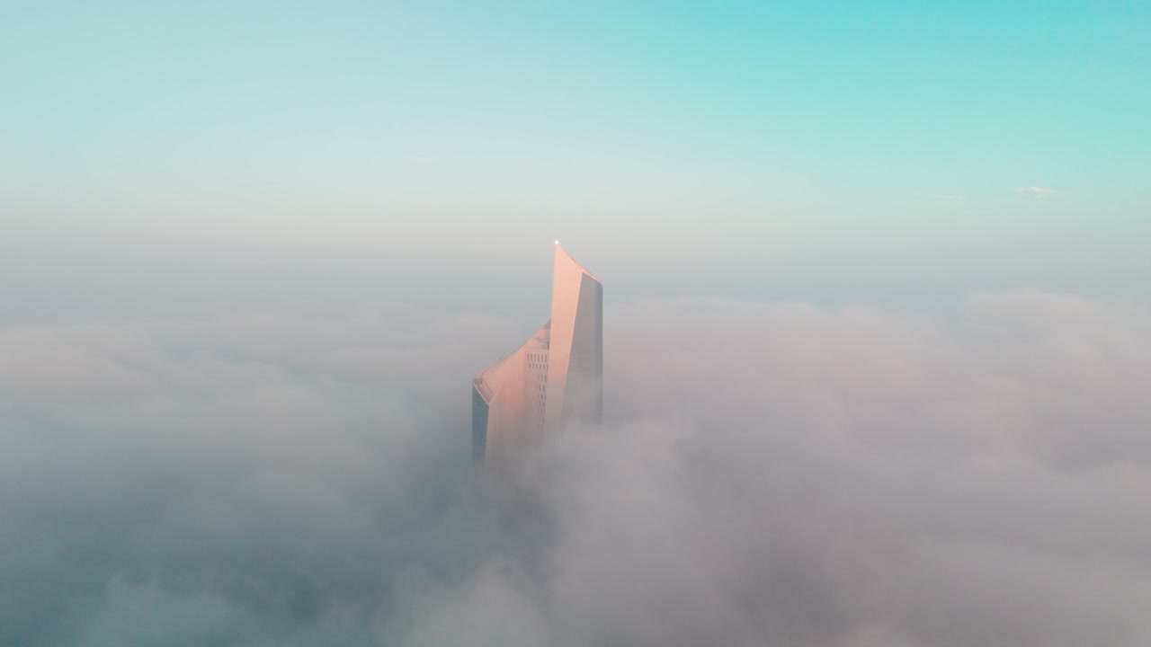 Cinematic drone aerial of a modern Kuwait City skyscraper piercing low clouds at sunrise, glowing in golden light with soft fog and blue sky—perfect for luxury, business, and futuristic skyline visual