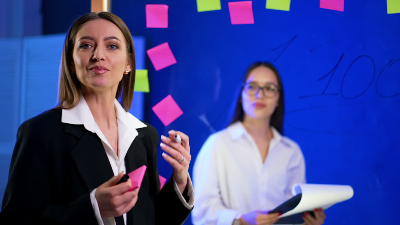 Positive outgoing active brunette talks near the glass wall with numbers and stickers on. Girl behind the board holds a clipboard.