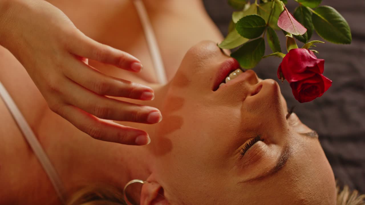 En attraktiv kvinde klædt i sexet lingeri leger forførende med en smuk rød rose, mens hun ligger i sengen