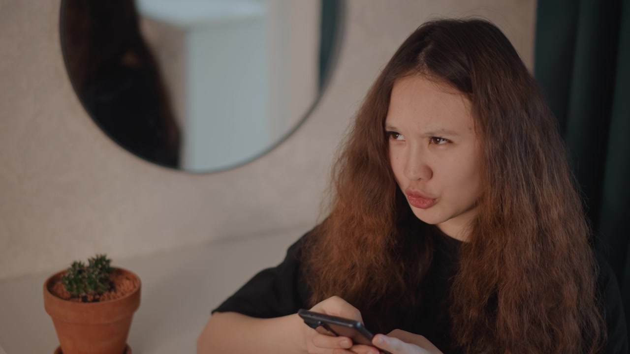 Young woman seated at vanity holds smartphone with both hands, reacting to screen with shifting expressions from focused reading to mild puzzlement and surprise while tapping and scrolling in soft