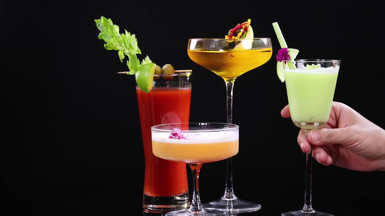 Vibrant cocktails in various glasses with garnishes, set against a dark backdrop, showcasing a hand holding one glass
