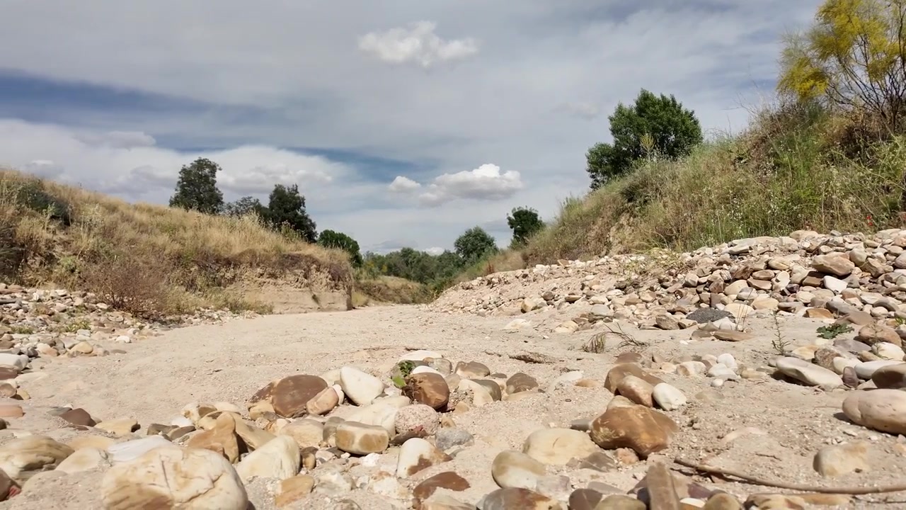 Ripresa a mano dal basso di un letto di fiume roccioso e prosciugato vicino a Madrid sotto il sole estivo. Terra arida, pietre sparse ed erbe dorate evocano l'impatto del cambiamento climatico e le condizioni di siccità persistente.