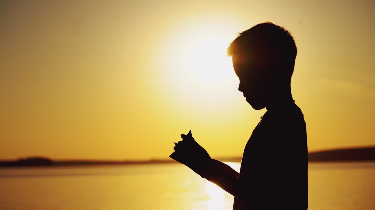 A little boy is making a paper boat with his own hands and playing with it in the air at sunset near the river in warm weather.
