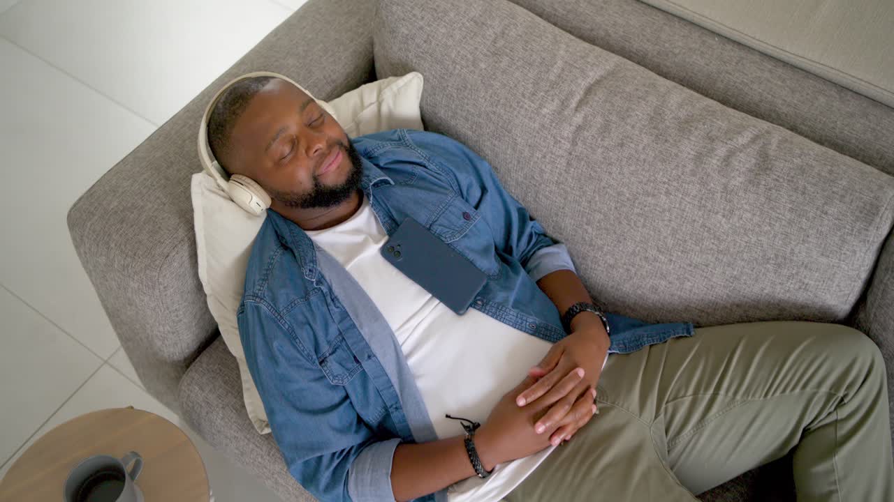Adult man holding smartphone tapping, resting phone on chest while relaxing with headphones on sofa