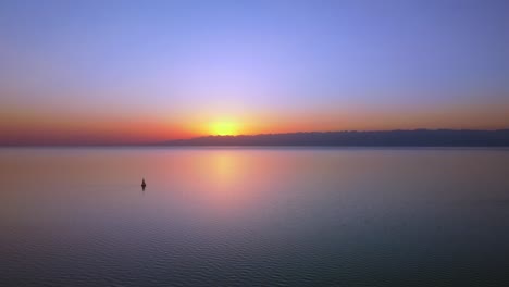 Luftaufnahmen-Eines-Großen-Sees,-Die-Während-Des-Sonnenuntergangs-Aufgenommen-Wurden,-Die-Lebendigen-Farben-Der-Untergehenden-Sonne,-Hoch-Aufragende-Berge-Im-Hintergrund-Und-Ein-Aktives-Segelboot-In-Der-Mitte