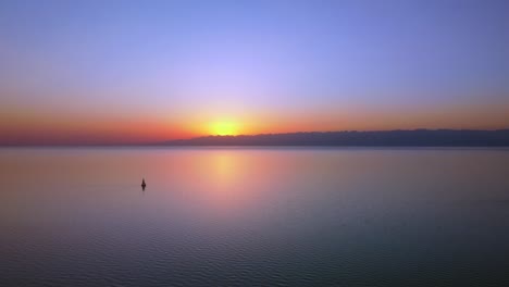 imágenes aéreas de un gran lago capturado durante la puesta de sol, los tonos vibrantes del sol poniente, imponentes montañas en el fondo y un velero activo en el centro