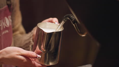 close up barista hands steaming milk inside stainless jug using espresso machine wand during beverage preparation, with soft lighting and warm tones