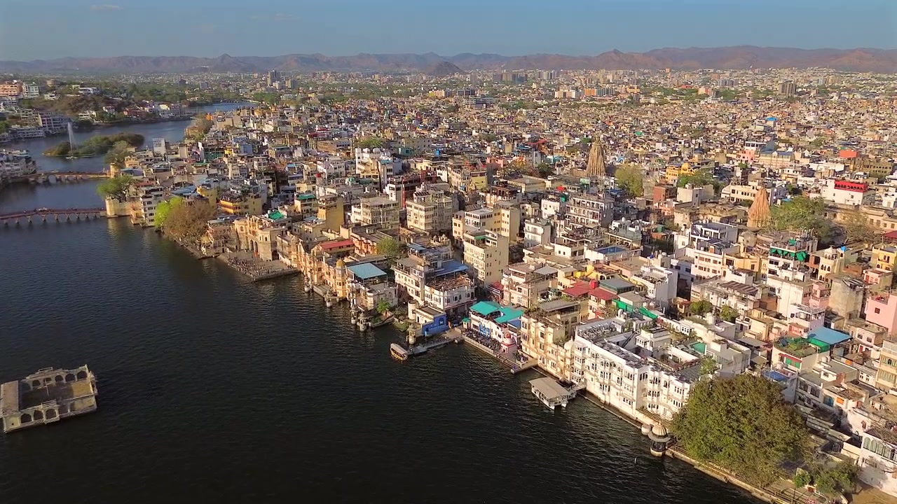 Pengambilan gambar statis sudut lebar dari ketinggian yang menampilkan lanskap perkotaan Udaipur yang semarak, dengan bangunan putih tradisional dan hotel warisan di sepanjang tepi Danau Pichola yang indah saat matahari terbenam, India