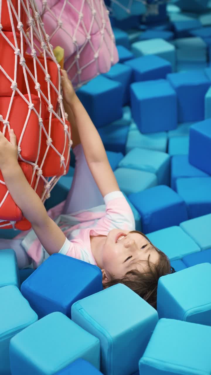 Happy young girl in striped dress lies back in a blue foam cube pit while holding onto a large hanging net bag filled with red foam blocks at an indoor playground