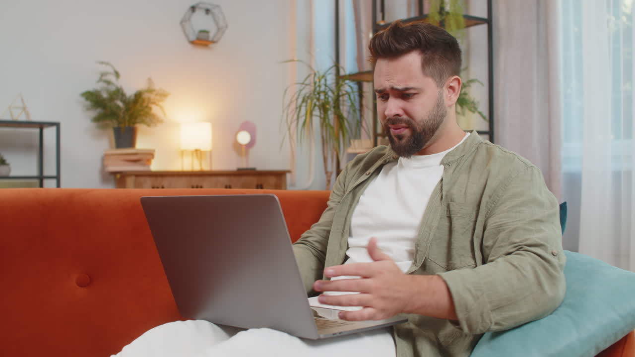 Upset young caucasian young man sitting on sofa at home using laptop having anxiety and stress