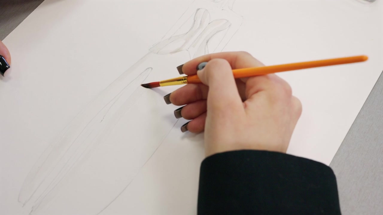A fashion design student begins a light pencil sketch of a female figure on white paper, with a watercolor palette and paint tray nearby