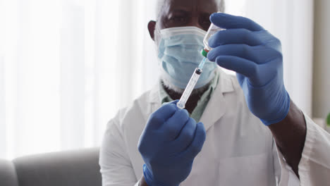 male african american doctor wearing face mask pulling out covid-19 vaccine into syringe