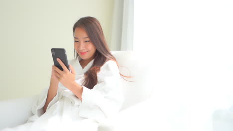 Lächelnde-Asiatische-Frau,-Die-Im-Sessel-Sitzt-Und-Den-Handybildschirm-Mit-Dem-Finger-Berührt-Und-Einen-Weißen-Hotelbademantel-Trägt