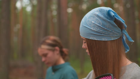 close-up of woman brushing her hair while in a serene forest setting, with a blue bandana tied around her head, in the blurred background, another woman focuses on opening her thermos