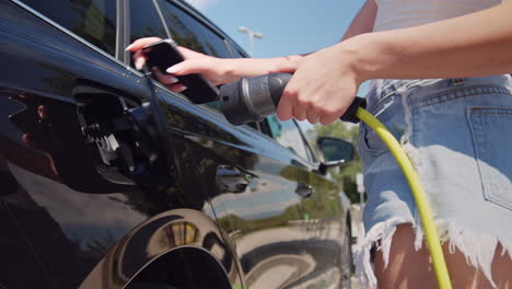 chica conectando un cargador a un coche eléctrico y usando un teléfono inteligente, de cerca