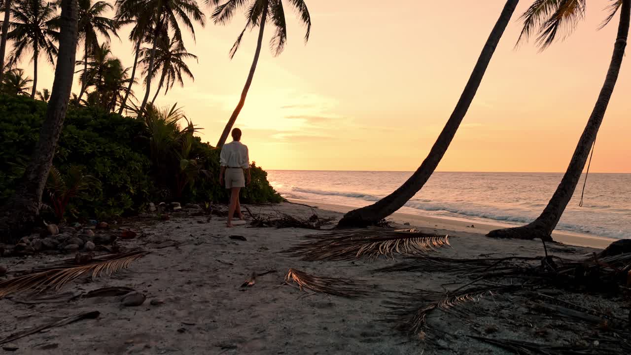 A stunning bird’s-eye perspective captures a lone woman standing barefoot on the white sands of Geiymiskih Fannu Beach as the Maldivian sun dips below the horizon.