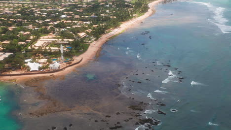 Aerial-view-of-Praia-do-Forte-beach,-the-coral-reef,-boats-parked,-palm-tree-area-on-a-cloudy-day,-Praia-do-Forte,-Bahia,-Brazil