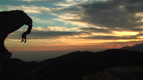 Panleft-De-Una-Silueta-Rockclimber-Colgando-De-Una-Pared-Rocosa-Que-Sobresale-Con-El-Atardecer-De-California-Más-Allá