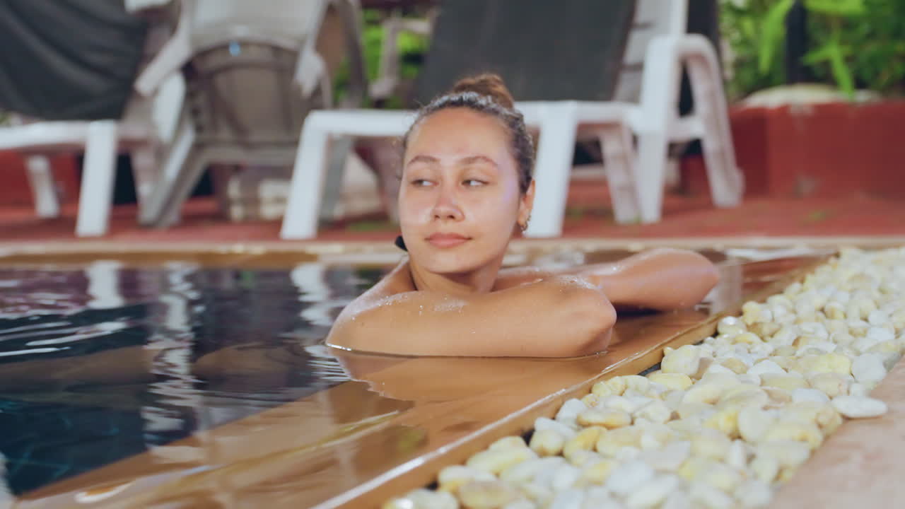Donna polinesiana che sogna ad occhi aperti a bordo piscina sull'isola, con il mento appoggiato su un bordo di legno con contorno di ciottoli, lettini visibili, luce solare soffusa, un'atmosfera da travel blogger, con un umore riflessivo e malinconico.