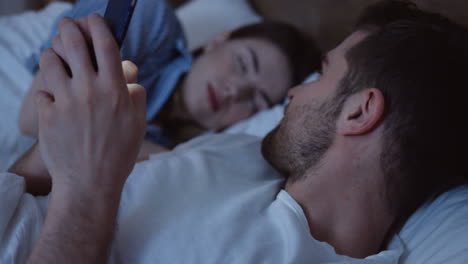 vista de cerca del joven caucásico acostado en la cama, charlando en el teléfono inteligente a altas horas de la noche y mirando a su esposa que duerme a su lado