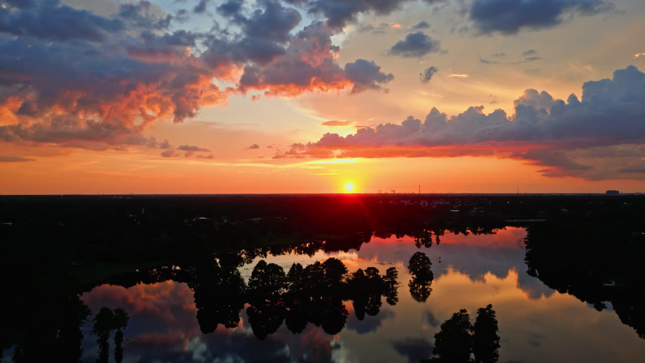 Brilliant orange and red sunset colors paint the sky over still water and forested edge, heavenly aerial backdrop background, aerial slowly orbiting to the left over the river