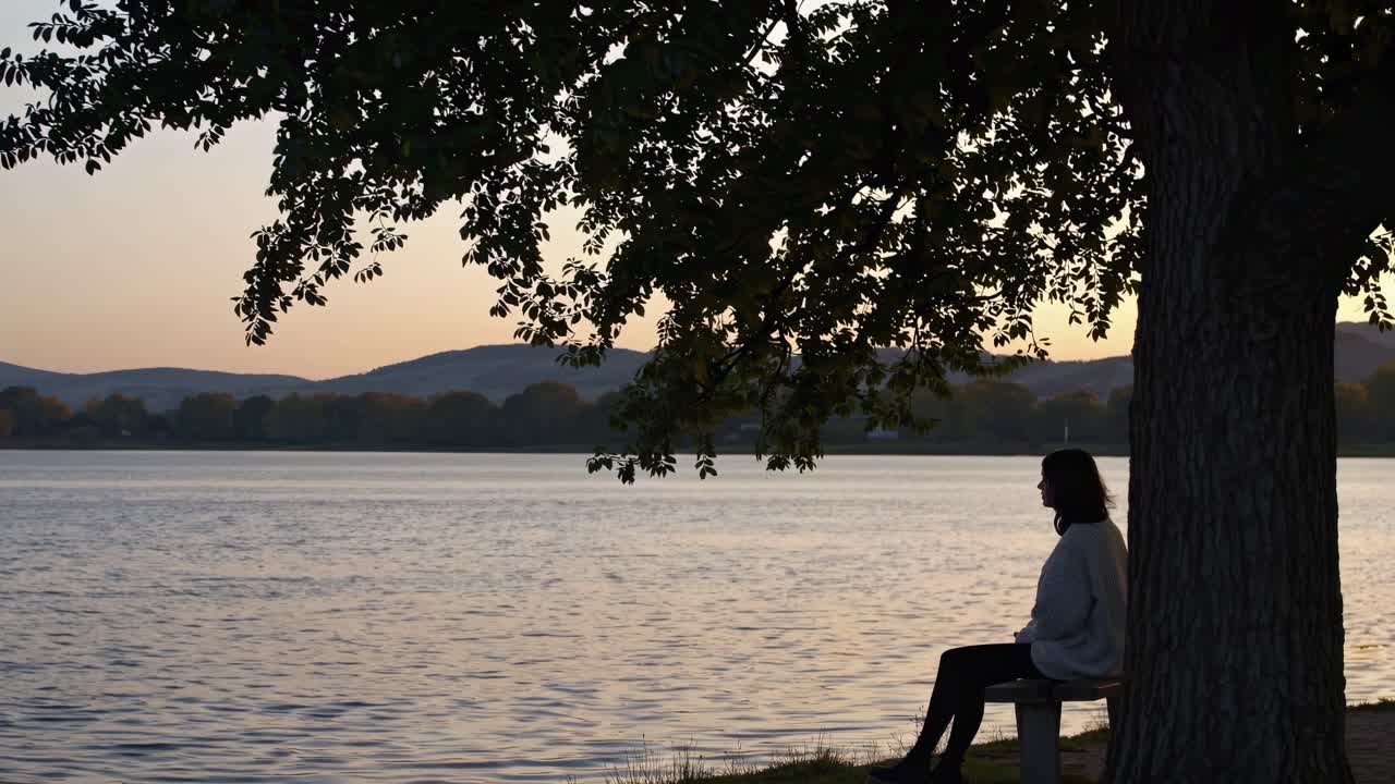 Silhouette di una persona seduta su una panchina in riva a un lago al tramonto, ripresa da un angolo laterale