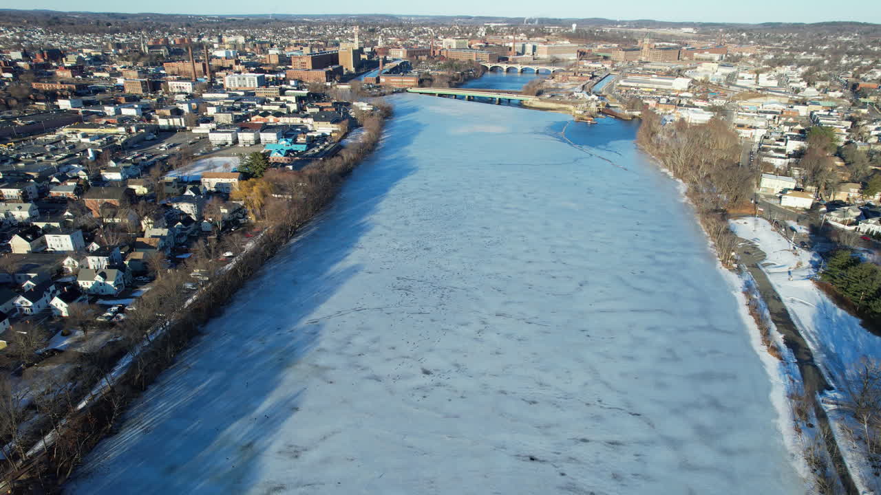 Aerial flyover frozen river and downtown of American town in Massachusetts. Sunny day in quiet American city of Lawrence. Icy and cold winter day in December. Wide shot.