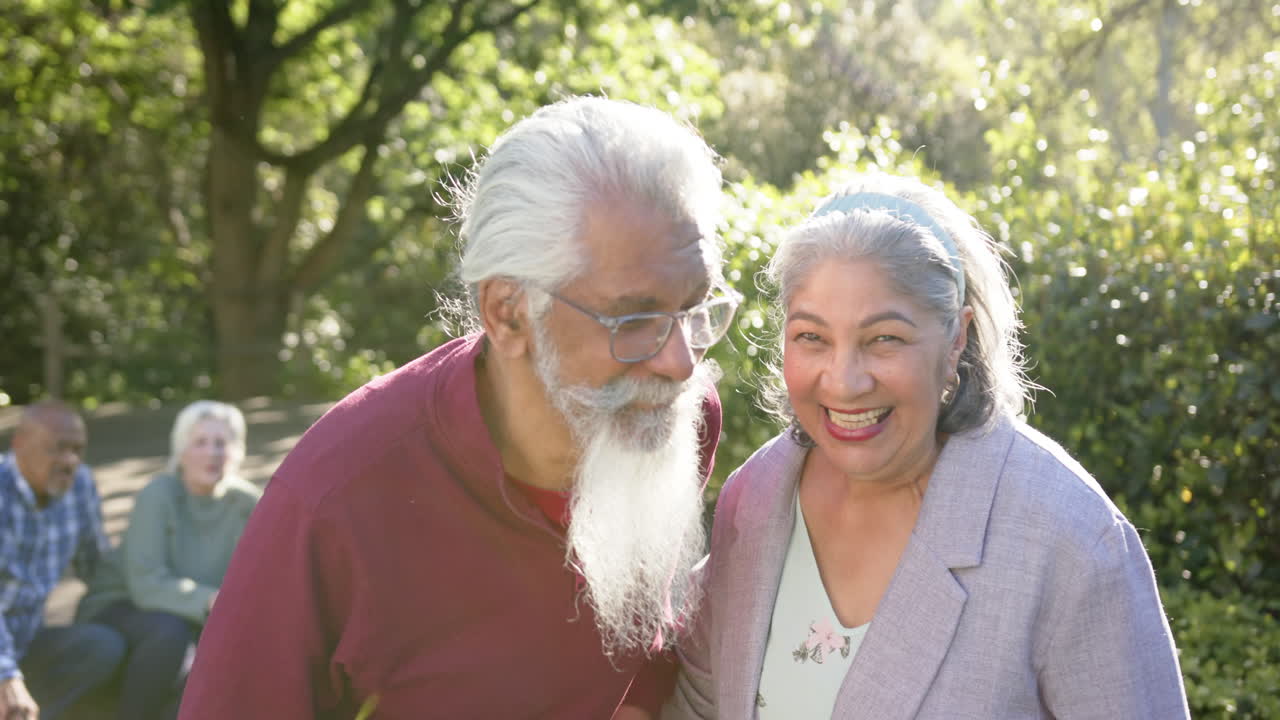 Retrato de feliz pareja mayor diversa abrazándose en un jardín soleado con amigos, a cámara lenta