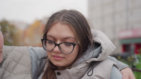 Close up of young woman seated outdoors resting her head on husband chest as he holds her closely, both dressed warmly, soft expression on her face, with blurred building