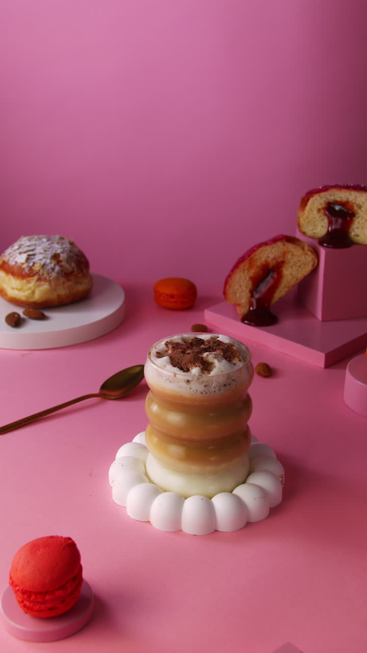 A feminine hand reaches to pick up a layered almond milk cappuccino in a curvy glass. The vibrant pink pastel setup features a jelly donut and macaron for a modern vegan café aesthetic