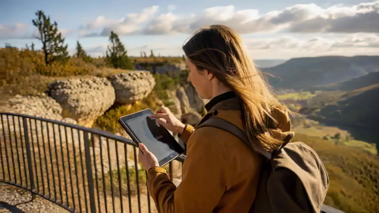 Reaching cliff-top viewpoint, woman hiker checking tablet and tapping stylus, brown jacket backpack