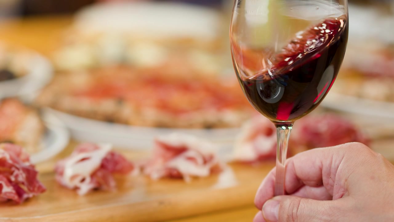 Una mano fa roteare vino rosso in un bicchiere su un tavolo con pizza e prosciutto