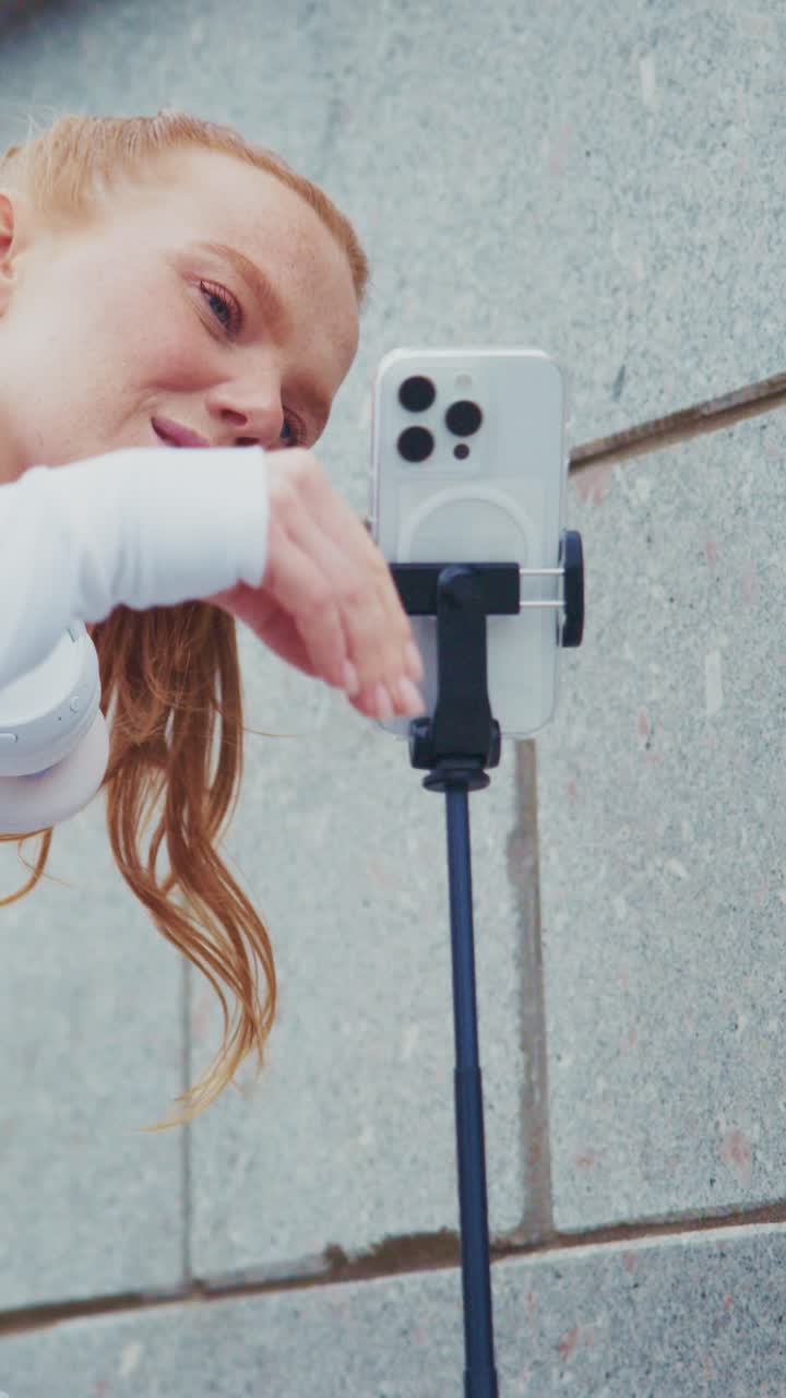A Young Woman Engaged in Taking Selfies with a Smartphone Attached to a Tripod, Capturing Her Expressions Against a Textured Wall Background